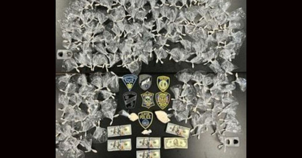 Dozens of small clear plastic bags containing white powdery substances arranged on a dark surface, alongside several police department patches and four $100 bills.