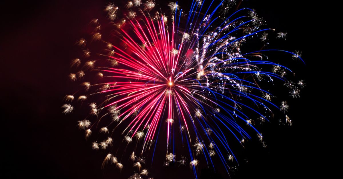 Red, white, and blue fireworks burst against a dark night sky during a Fourth of July celebration.