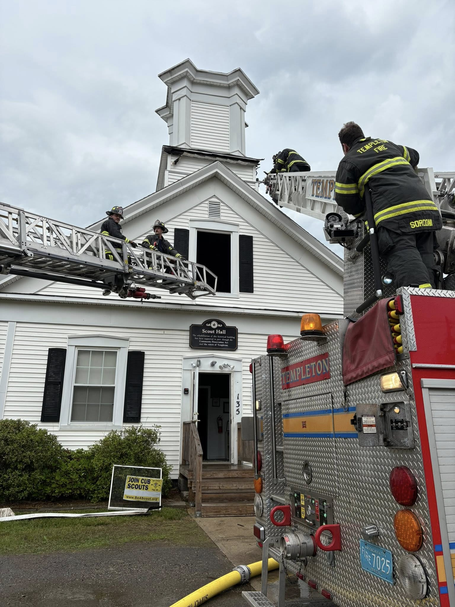 Firefighters on ladder trucks inspect the damaged cupola of a white building labeled “Scout Hall” after a lightning strike caused a fire. A Templeton fire truck is parked in front.