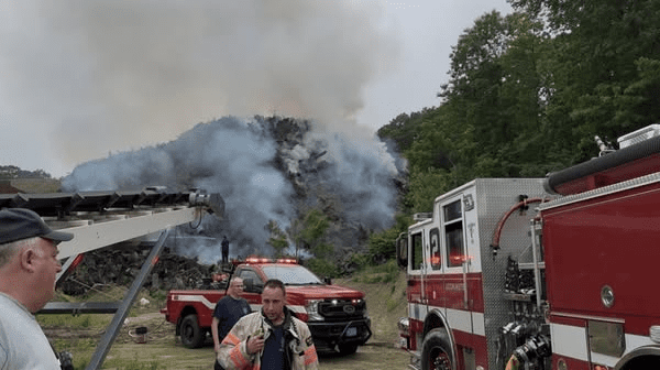 Firefighters and fire trucks at the scene of a large brush fire with heavy smoke rising from a pile of burning stumps near a wooded area.