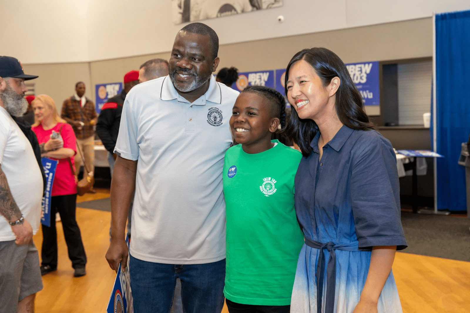 Mayor Michelle Wu stands smiling with Josué Pierre, who is wearing a light gray IBEW Local 103 polo, and a child in a green IBEW shirt at an indoor union event with supporters in the background.
