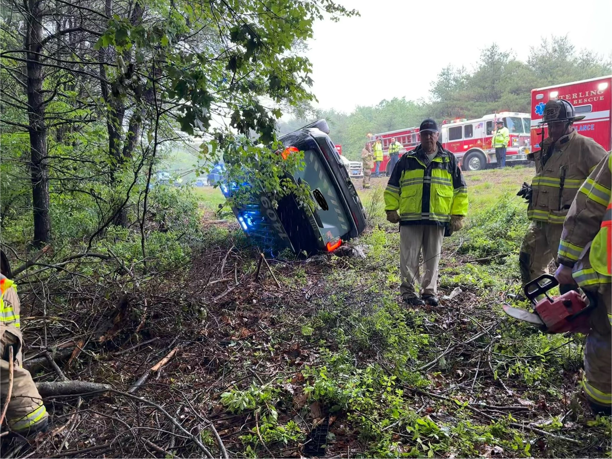 Firefighters surround an overturned State Police cruiser in a wooded area off I-190 in Sterling, with emergency vehicles and personnel visible in the background.