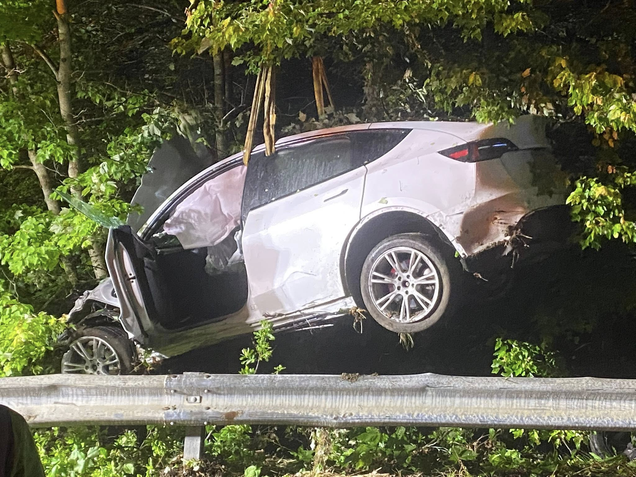 A damaged white vehicle being hoisted from a wooded area beside the Massachusetts Turnpike at night, with airbags deployed and vegetation entangled around the vehicle.