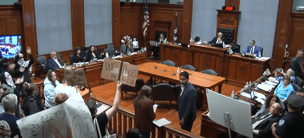 Wide view of Worcester City Council chamber: councillors sit at the dais while a crowd of protesters at left hold cardboard and poster-board signs—one reads “DUE PROCESS FOR ALL”—and shout toward the officials.