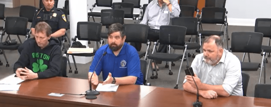 Three men seated at a table during the June 10, 2025 Leominster Legal Affairs Committee meeting. From left to right: Kevin Crowley, owner of 25 Mohawk Drive, wearing a black Boston Celtics hoodie; Luke Beaulac, City Code Enforcement Officer, in a blue shirt speaking into a microphone; and a representative from Apical, wearing a light-colored shirt, holding a phone. Several empty chairs and a uniformed police officer are visible in the background.