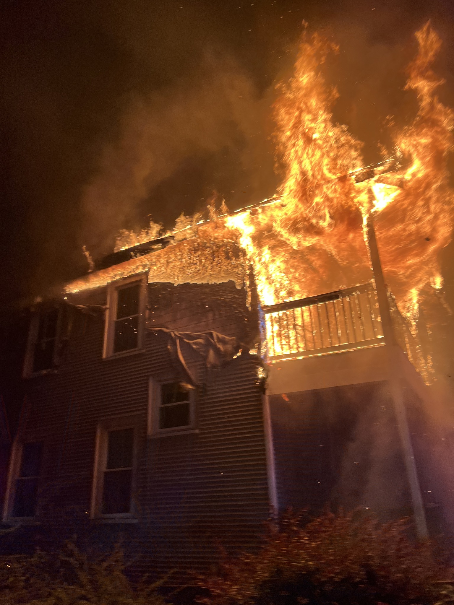 A nighttime image of a house fire showing intense flames consuming the roof and second floor of a two-story home, with smoke and embers filling the sky. (Photo Credit: Framingham Fire Department)
