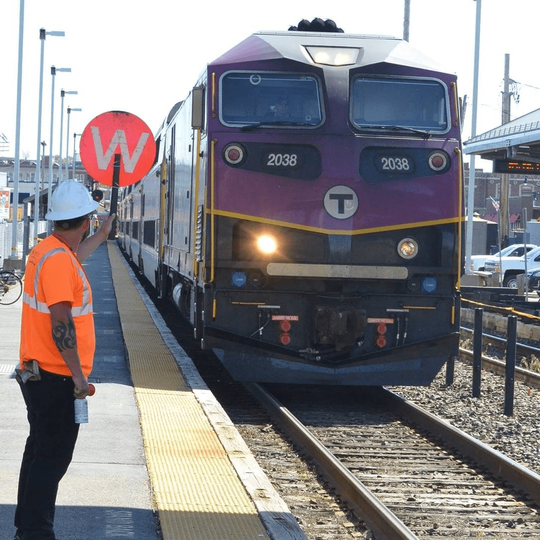 Keolis worker in orange shirt and white hard hat signals westbound MBTA train 1503 with a red "W" sign at Framingham Station; train 2038 approaches on track 1 during track maintenance.